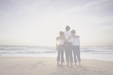 Image of a family of three with arms around each other at the beach shore facing away looking out over the water